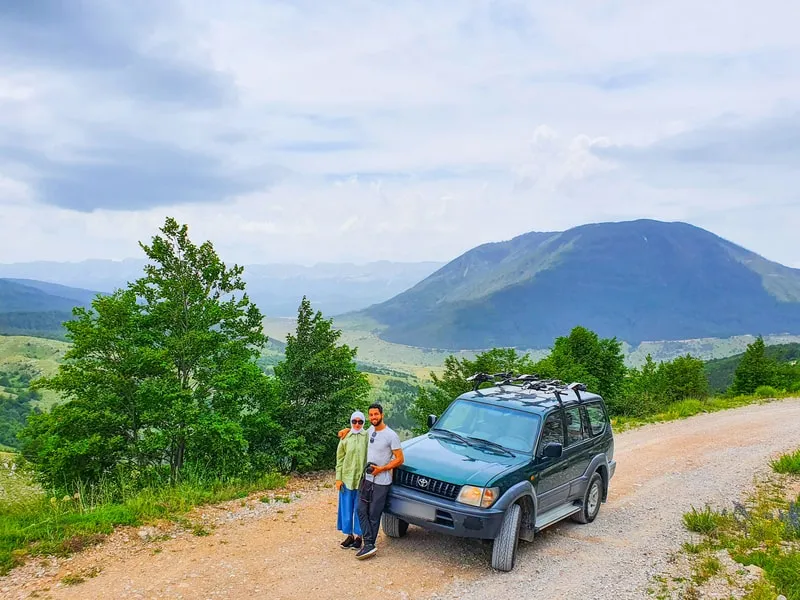 Two travellers beside the 4x4 on a forest track, with a mountain behind