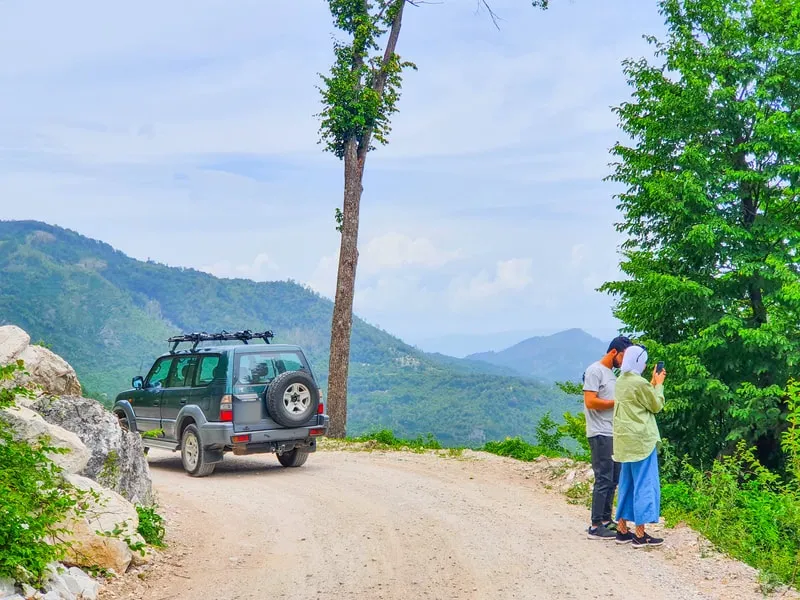 Two travellers looking out over wooded valleys, the 4x4 parked behind them